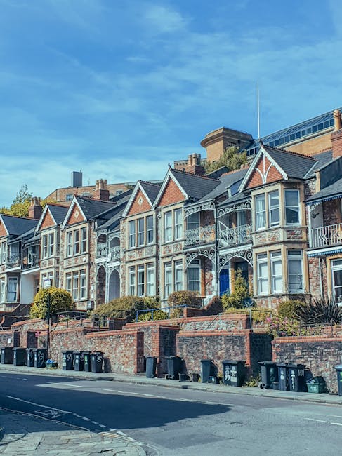 A row of Victorian-style terraced houses with decorative cast iron balconies and bay windows, situated on a sloped street with brick front gardens and retaining walls. The houses are multi-storey, with brick and painted facades, and have roofs covered with dark tiles. Below the houses, black wheelie bins are lined up along the pavement, and the street is paved with asphalt. The scene is captured on a clear day with a bright blue sky and some wispy clouds, suggesting good weather for home relocation and furniture transport. The image highlights a typical residential area suitable for house removal services, with no vehicles or moving equipment visible in the scene but fitting for the context of furniture removals for Finsbury Park flats, as offered by Man and Van Finsbury Park.