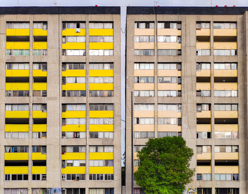 Detailed exterior view of a multi-storey apartment block with concrete facade, featuring numerous windows and predominantly yellow horizontal balcony railings on the left-side building. The right-side section has similar windows with beige and white panels, some with visible curtains or blinds. A large green tree is situated at the base of the buildings, partially obscuring the lower floors. The sky is visible at the top of the image, with the buildings occupying most of the frame. This residential setting suggests urban living spaces that may be involved in home relocation or furniture transport activities. The scene is well-lit with natural daylight, emphasizing the structural details and the contrasting colors of the balconies and building facades, supporting visual context for house removals or moving services at properties like those on Andover Estate, Finsbury Park.