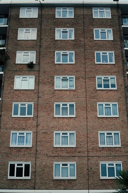 A row of Victorian-style terraced houses with decorative cast iron balconies and bay windows, situated on a sloped street with brick front gardens and retaining walls. The houses are multi-storey, with brick and painted facades, and have roofs covered with dark tiles. Below the houses, black wheelie bins are lined up along the pavement, and the street is paved with asphalt. The scene is captured on a clear day with a bright blue sky and some wispy clouds, suggesting good weather for home relocation and furniture transport. The image highlights a typical residential area suitable for house removal services, with no vehicles or moving equipment visible in the scene but fitting for the context of furniture removals for Finsbury Park flats, as offered by Man and Van Finsbury Park.