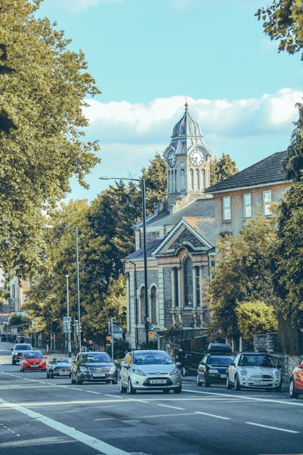 A street scene in Finsbury Park featuring a historic building with a clock tower, surrounded by green trees and residential and commercial properties. Several cars are parked along the roadside, and others are driving on the road, which is marked with lane lines and pedestrian crossings. The sky is blue with some scattered clouds. The area appears busy with typical urban activity, supporting the context of home relocation and moving logistics. This image aligns with the services offered by Man and Van Finsbury Park, highlighting the environment where furniture transport and packing and moving activities might take place.