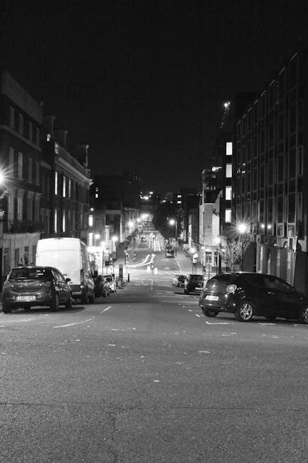 A nighttime street scene showing a residential and commercial area with several parked cars along the curb, including a white van and a black SUV positioned close to the sidewalk, indicating active loading or unloading. The road is lined with multi-storey buildings, some with illuminated windows and exterior lights, creating a contrast against the dark sky. In the distance, the street extends towards a slight incline, with streetlights and traffic signals visible at the intersection. The pavement appears clear, and the overall environment suggests a quiet urban setting suitable for home relocations or furniture transport. This scene illustrates the typical environment where Man and Van Finsbury Park provides house removals and moving logistics, involving packing, moving, and loading procedures.
