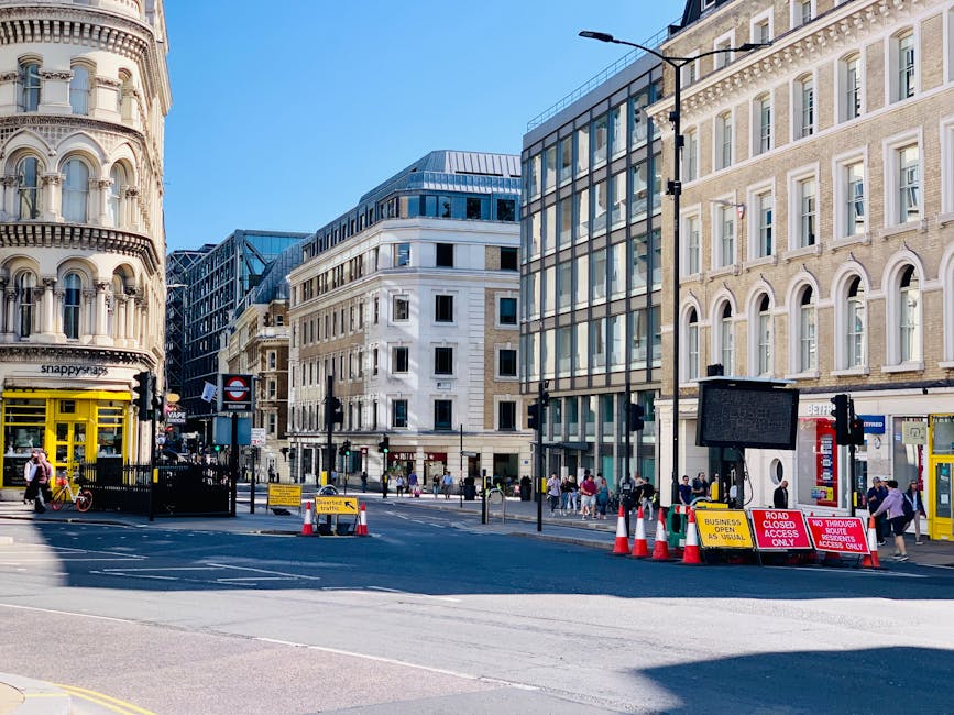 A busy urban street scene in Finsbury Park with a mix of modern and historic buildings, including a rounded corner building on the left and a glass-fronted office building on the right. Pedestrians are walking on the sidewalks, some crossing the road, while a temporary pedestrian access gate is visible blocking part of the street. Several traffic cones and road closure signs are set up at the intersection, indicating roadwork or restricted access, with signs mentioning 'business access only' and 'road closed.' The sky is clear and sunny, with a black lamppost and a digital information board also visible, suggesting an active area with ongoing logistics or transportation activity, relevant to house and office removals or relocation services.