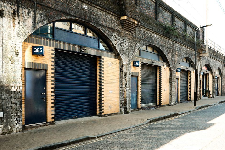 A row of industrial-style commercial units with large, dark blue metal roller shutter doors and brick facades featuring black brick accents around each entrance, numbered 385, 386, 387, and 388, situated along a paved street in an urban setting. The units are built beneath a curved brick railway arch with weathered bricks and a small window beneath the arch. A streetlamp is positioned in front of the units, and the sidewalk shows signs of light wear. This scene illustrates potential locations for house or office removals, where items are loaded or unloaded from vehicles such as vans or trucks. The image is captured during daylight with clear shadows, exemplifying a typical commercial frontage suitable for professional relocation or furniture transport services offered by companies like Man and Van Finsbury Park, highlighting packing, loading, and logistics activities in urban environments.