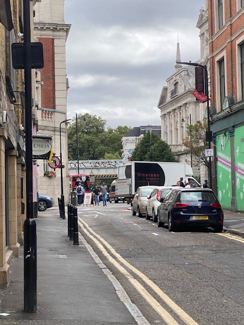 A busy urban street scene in Finsbury Park with a mix of modern and historic buildings, including a rounded corner building on the left and a glass-fronted office building on the right. Pedestrians are walking on the sidewalks, some crossing the road, while a temporary pedestrian access gate is visible blocking part of the street. Several traffic cones and road closure signs are set up at the intersection, indicating roadwork or restricted access, with signs mentioning 'business access only' and 'road closed.' The sky is clear and sunny, with a black lamppost and a digital information board also visible, suggesting an active area with ongoing logistics or transportation activity, relevant to house and office removals or relocation services.