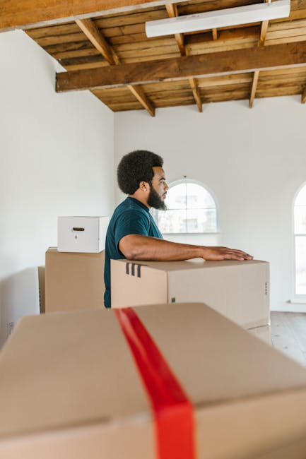 A man with a dark beard and curly hair, dressed in a blue T-shirt, is shown standing inside a well-lit room with white walls and a wooden ceiling. He is positioned behind a large cardboard box, with his hands resting on top, preparing for or during a home relocation. Several other cardboard boxes are stacked and placed across the room, some with red packing straps. The room features two arched windows, allowing natural daylight to brighten the space. The setting suggests an ongoing furniture transport or packing process, typical of removals or moving services like those provided by Man and Van Finsbury Park. The environment indicates an organized loading or unpacking scene, emphasizing aspects of packing and moving logistics within a home interior.