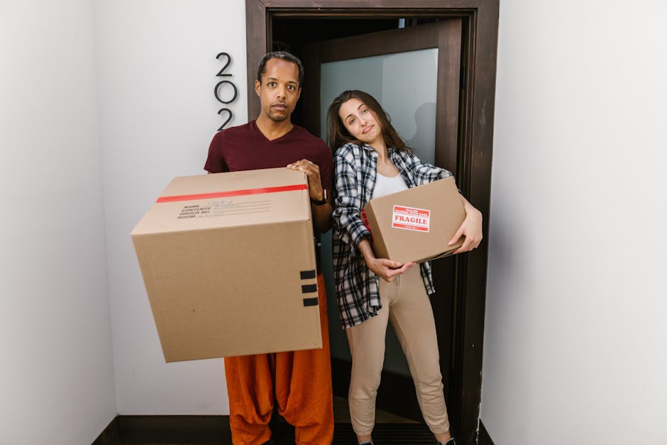 A man and a woman stand inside a doorway with a dark wooden frame, each holding cardboard boxes filled with household items, preparing for a home relocation. The man, wearing a maroon T-shirt and orange trousers, holds a large box with visible black tape and red markings, while the woman, dressed in a plaid shirt and beige trousers, carries a smaller box labeled 'Fragile' with red and white tape. Behind them, to the left of the door, is the house number '200.' The scene is set inside the entrance area, with smooth white walls and a dark baseboard visible at the bottom, indicating a clean, modern interior. The woman leans slightly against the doorframe with a relaxed expression, and the man looks directly at the camera with a neutral expression. This image captures the process of packing and loading items during furniture transport with evident attention to careful handling of fragile belongings, typical of professional removals by Man and Van Finsbury Park, in preparation for house moving at Finsbury Park Station N4.